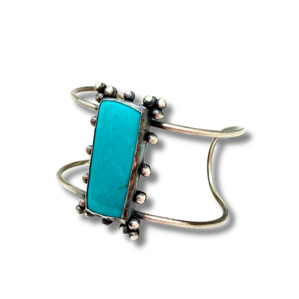 A silver cuff bracelet featuring a large, polished turquoise rectangular stone in a bezel setting with small silver beads along the border, shown on a white background with a soft drop shadow.
