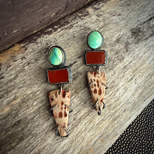 A pair of handmade dangle earrings displayed on a rustic weathered wood surface. The earrings feature a round turquoise cabochon, a rectangular carnelian stone, and a tapered fossil wood piece with distinctive brown spots.