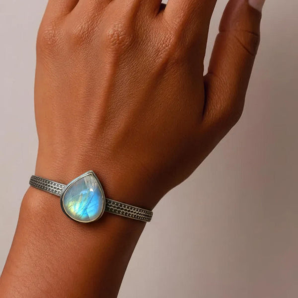 A close-up of a person's wrist wearing a silver-toned bangle bracelet with a teardrop-shaped iridescent moonstone cabochon.
