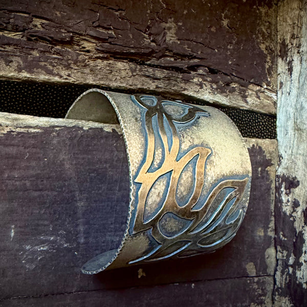 A close-up of a wide oxidized silver cuff bracelet with an embellished leaf-like pattern, resting against a dark rustic wooden surface.