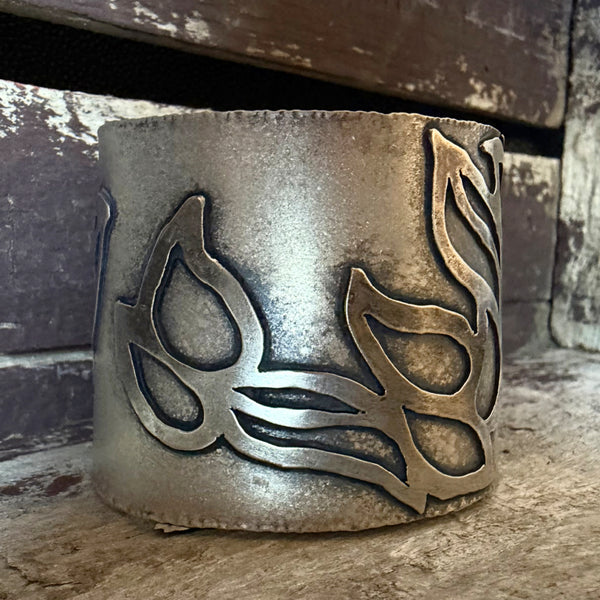 A close-up of a wide oxidized silver cuff bracelet with an embellished leaf-like pattern, resting against a dark rustic wooden surface.