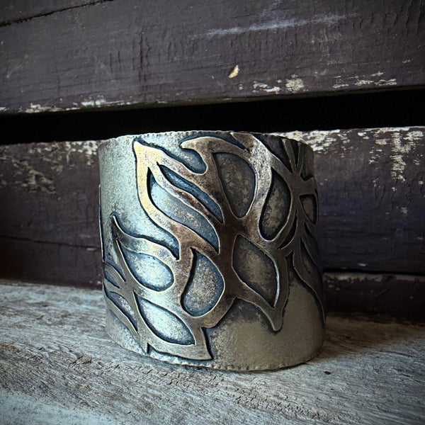 A close-up of a wide oxidized silver cuff bracelet with an embellished leaf-like pattern, resting against a dark rustic wooden surface.