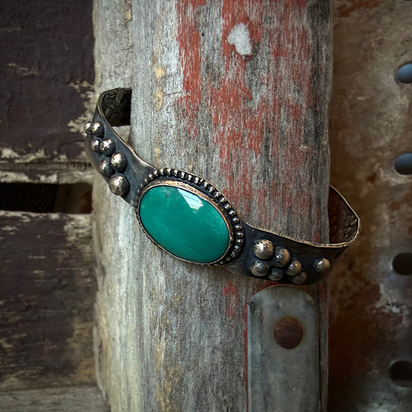 An oxidized silver cuff bracelet with an oval turquoise stone and decorative bead details, displayed on a rustic dark wooden surface.