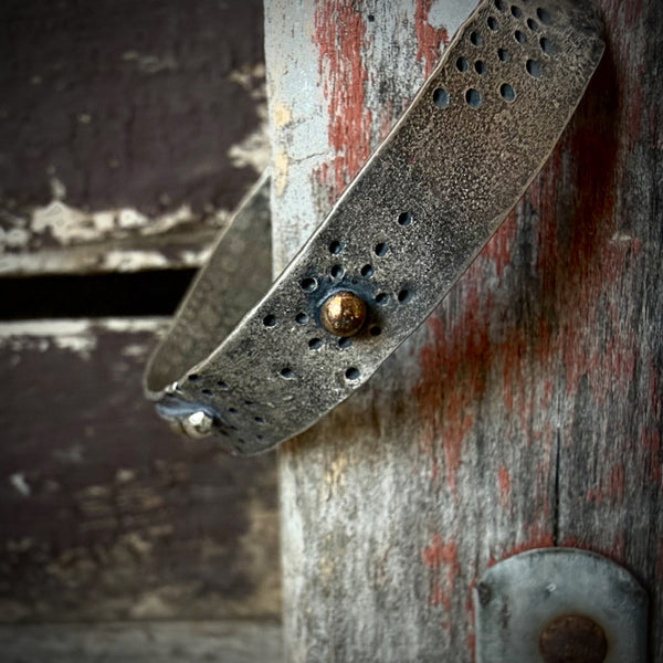 A close-up of a rustic metal cuff bracelet with a hammered texture, punched holes, and a central bronze gall, resting against a weathered wooden post with peeling red paint.