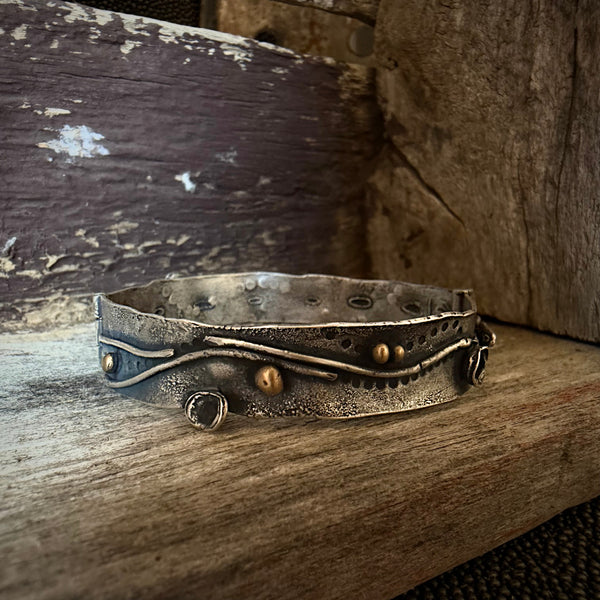 A handmade silver cuff bracelet with organic wavy lines, small bronze beads, and a spiral detail, resting on a rustic wooden surface.