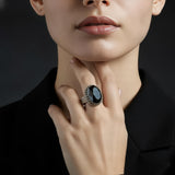 A close-up lifestyle shot of a model wearing a large silver ring with a dark blue oval cabochon stone, set against a dark background.