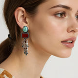 A close-up of a woman wearing a long drop earring with a turquoise oval stone, a red rectangular stone, and fossil stones, paired with a cream and brown patterned top against a light gray background.