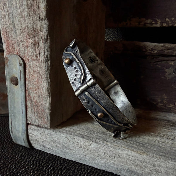 A handmade metal cuff bracelet with bronze accents and a hinge closure, resting on a rustic wooden surface.