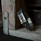 A rustic metal cuff bracelet with a decorative buckle and bronze accents, resting on weathered wood next to a metal hinge.