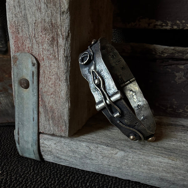 A rustic metal cuff bracelet with a decorative buckle and bronze accents, resting on weathered wood next to a metal hinge.