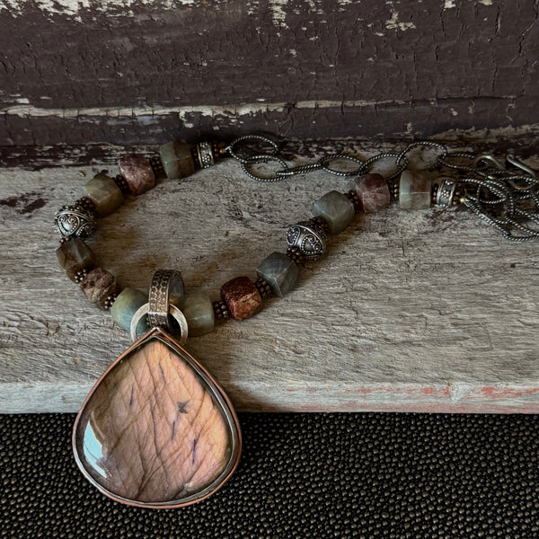 A necklace with a pink labradorite pendant and earth tone square sapphire stones lays on a wood background