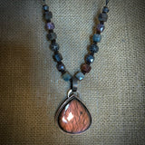 A beaded necklace featuring a large teardrop-shaped pendant made of mauve labradorite stone, suspended from dark metal and earth tone beads on a textured burlap background.