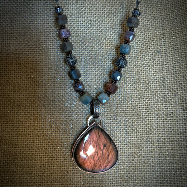 A beaded necklace featuring a large teardrop-shaped pendant made of mauve labradorite stone, suspended from dark metal and earth tone beads on a textured burlap background.