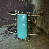 A silver cuff bracelet featuring a large, polished turquoise rectangular stone in a beaded border setting, displayed on a rustic wooden surface against a weathered wood background.