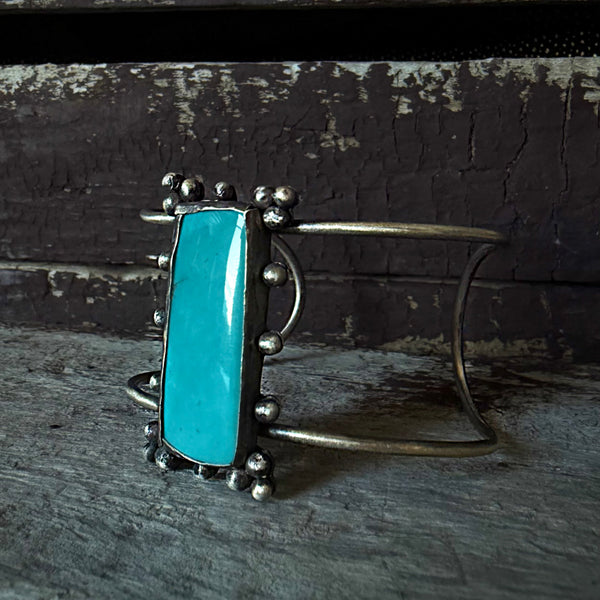 A sterling silver cuff bracelet featuring a large, vibrant turquoise rectangular stone in a beaded border setting, displayed on a rustic weathered wooden surface.