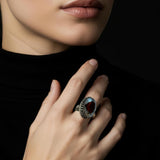 A close-up of a model's hand wearing a silver ring with a large oval Blue gemstone, set against a dark black background.
