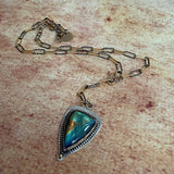A silver and gold chain necklace with a triangular pendant featuring a blue-green labradorite stone, displayed on a textured beige surface.