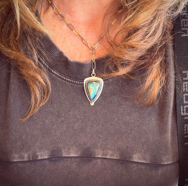 A close-up of a person wearing a silver and gold necklace with a triangular pendant featuring a blue and green labradorite stone, styled over a dark gray shirt with blonde hair framing the neck.