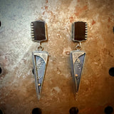 A pair of handmade silver dangle earrings with black onyx rectangular top sections and long pointed silver drops, displayed on a textured, rustic brown background.