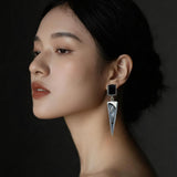 A close-up side profile of a woman wearing a long, geometric silver dangle earring with a black rectangular top and a silver leaf-shaped drop featuring engraved details.
