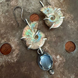 A pair of handmade dangle earrings featuring natural ammonite shells with iridescent green and blue patina, accented with small silver prongs, and a silver-toned flower-shaped pendant with a blue moonstone, displayed on a rustic metal surface.