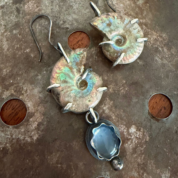 A pair of handmade dangle earrings featuring natural ammonite shells with iridescent green and blue patina, accented with small silver prongs, and a silver-toned flower-shaped pendant with a blue moonstone, displayed on a rustic metal surface.