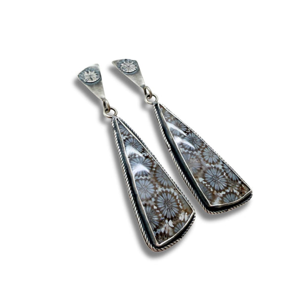 A pair of silver dangle earrings featuring large, teardrop-shaped black fossil coral pendants with a detailed floral or spiderweb-like pattern in gray, white, and brown, set in a twisted rope-style silver border.
