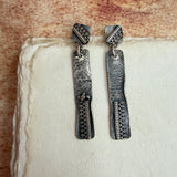 A pair of handmade silver dangle earrings with textured, hammered rectangular drops and geometric engraved details, displayed on a piece of textured off-white paper against a warm brown background.
