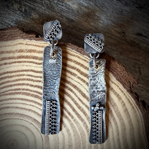 A pair of handmade silver dangle earrings with textured, hammered rectangular drops and geometric engraved details, displayed on a piece of textured off-white paper against a warm brown background.