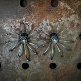 Two matching metallic starburst-style earrings with multiple thin radiating arms, shown on a distressed rusty metal surface.