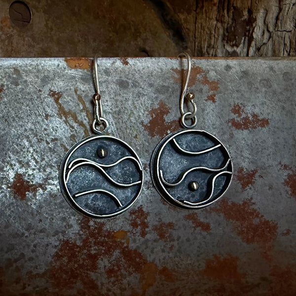 A pair of handmade silver dangle earrings with circular wave designs, displayed on a rustic, weathered metal surface.