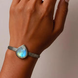 A close-up of a person's wrist wearing a silver-toned bangle bracelet with a teardrop-shaped iridescent moonstone cabochon.