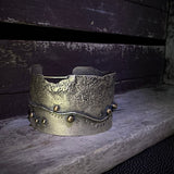 A distressed silver metal cuff bracelet with a textured, hammered finish and small bronze embellishments, displayed on a weathered wooden surface.