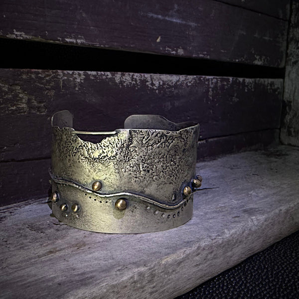 A distressed silver metal cuff bracelet with a textured, hammered finish and small bronze embellishments, displayed on a weathered wooden surface.