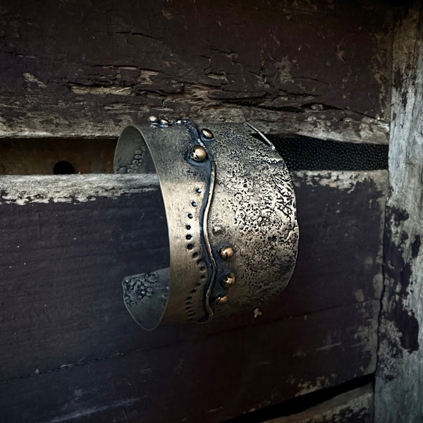 A distressed silver metal cuff bracelet with a textured, hammered finish and small bronze embellishments, displayed on a weathered wooden surface.