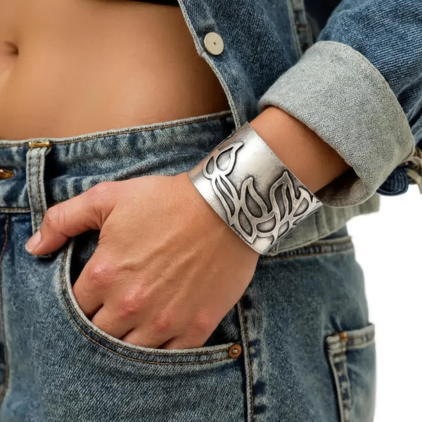 A close-up of a person wearing a wide silver cuff bracelet with engraved abstract patterns, paired with a denim jacket and blue jeans.