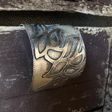 A close-up of a wide oxidized silver cuff bracelet with an embellished leaf-like pattern, resting against a dark rustic wooden surface.