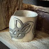 A close-up of a wide oxidized silver cuff bracelet with an embellished leaf-like pattern, resting against a dark rustic wooden surface.
