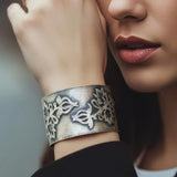 A close-up of a woman's wrist wearing a wide silver cuff bracelet with intricate floral cutout patterns, against a soft blurred background.