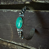 An oxidized silver cuff bracelet with an oval turquoise stone and decorative bead details, displayed on a rustic dark wooden surface.