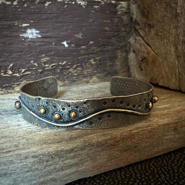 A handmade silver cuff bracelet with a wavy center line and small bronze balls, displayed on a rustic wooden surface.
