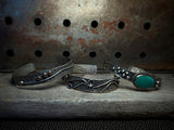 Three handmade silver cuff bracelets with rustic textures and accents, including one with a prominent turquoise stone, displayed on a weathered wooden surface against a rusty metal background.