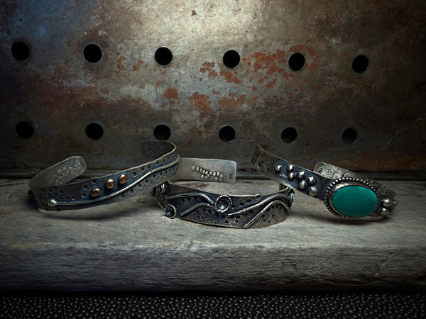 Three handmade silver cuff bracelets with rustic textures and accents, including one with a prominent turquoise stone, displayed on a weathered wooden surface against a rusty metal background.