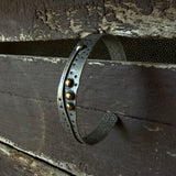 A rustic metal cuff bracelet with a weathered dark finish, featuring a central channel with circular cutouts, three bronze balls, and a textured industrial design, shown wrapped around weathered wooden planks.