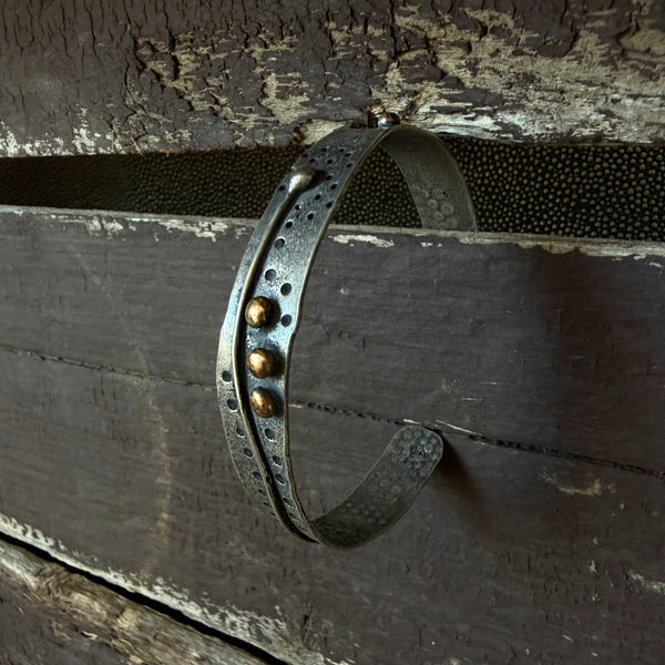 A rustic metal cuff bracelet with a weathered dark finish, featuring a central channel with circular cutouts, three bronze balls, and a textured industrial design, shown wrapped around weathered wooden planks.