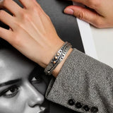 A close-up of a person's wrist wearing a textured silver cuff bracelet with three small round pearl-like accents, paired with a gray patterned blazer. A black-and-white portrait print and another hand are visible in the background.