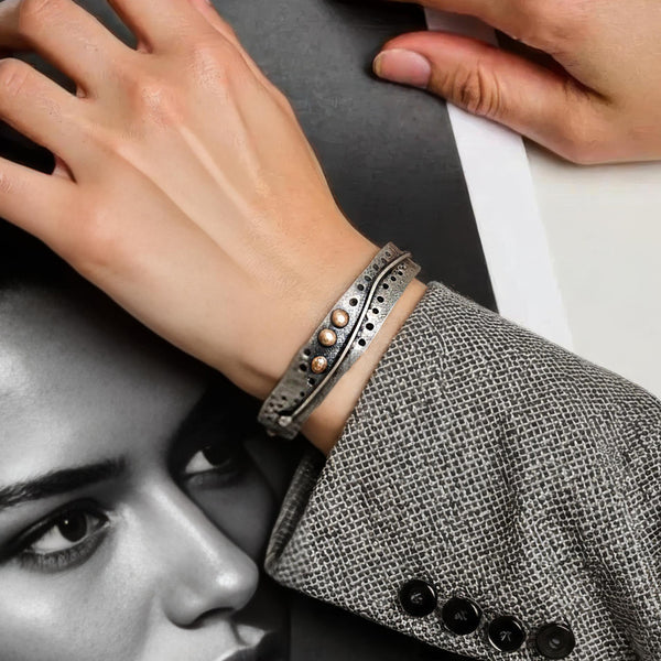A close-up of a person's wrist wearing a textured silver cuff bracelet with three small round pearl-like accents, paired with a gray patterned blazer. A black-and-white portrait print and another hand are visible in the background.