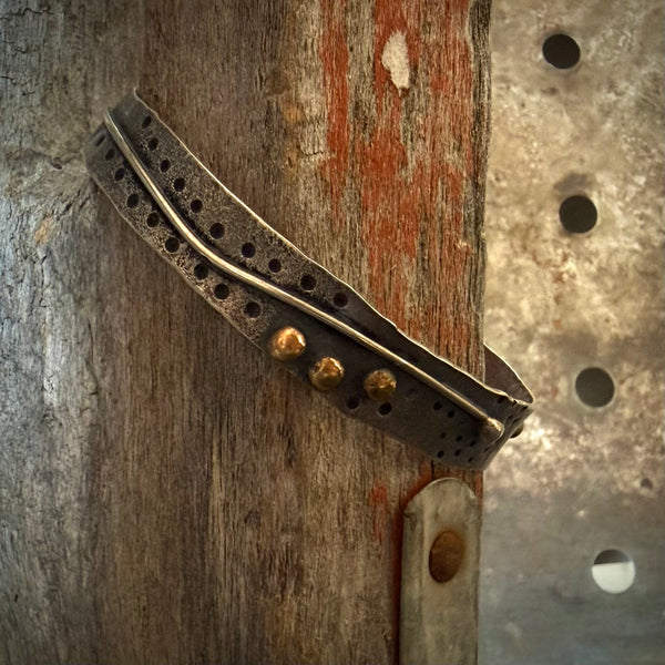 A rustic metal cuff bracelet with a weathered dark finish, featuring a central channel with circular cutouts, three bronze balls, and a textured industrial design, shown wrapped around a weathered wooden post.
