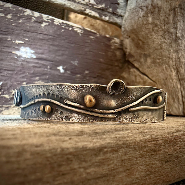 A handmade silver cuff bracelet with organic wavy lines and small bronze bead accents, resting on a rustic wooden surface.