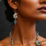 A close-up of a model wearing a chunky silver-toned necklace with a curved white feather centerpiece, black and blue stone accents, and matching dangling earrings against a dark gray background.